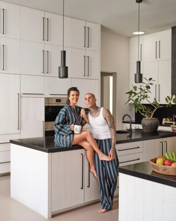 Kourtney Kardashian and Travis Barker pose in a modern kitchen. Kourtney sits on a kitchen island next to Barker.  