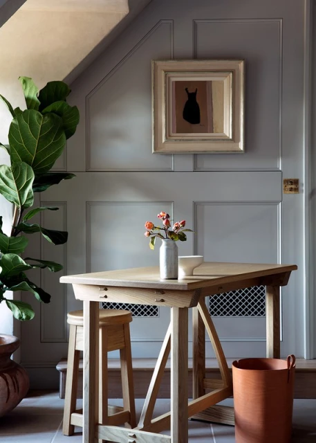 Interior setting showing a light wood table set with a vase of flowers, stool, and framed art against a grey panelled wall.