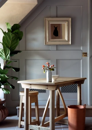 Interior setting showing a light wood table set with a vase of flowers, stool, and framed art against a grey panelled wall.