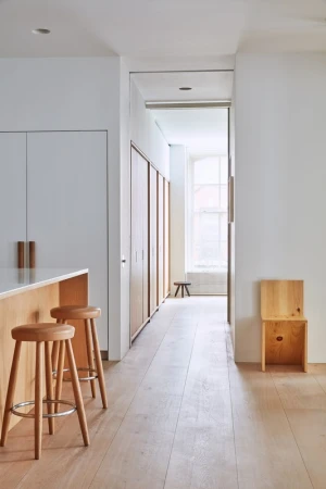Minimalist interior hallway featuring light wood flooring, wooden bar stools, and a bright window at the far end
