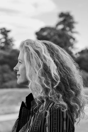 Profile portrait of a woman with long curly hair wearing a striped patterned jacket against an outdoor landscape background.