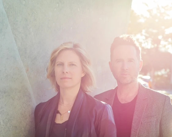 Portrait of a blonde woman and a dark-haired man standing together outdoors with warm sunlight flaring across the upper right frame.
