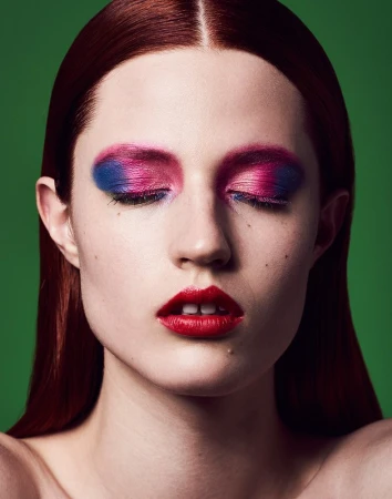 Close-up portrait of a woman with vibrant pink and blue eyeshadow and bold red lipstick against a solid green background.