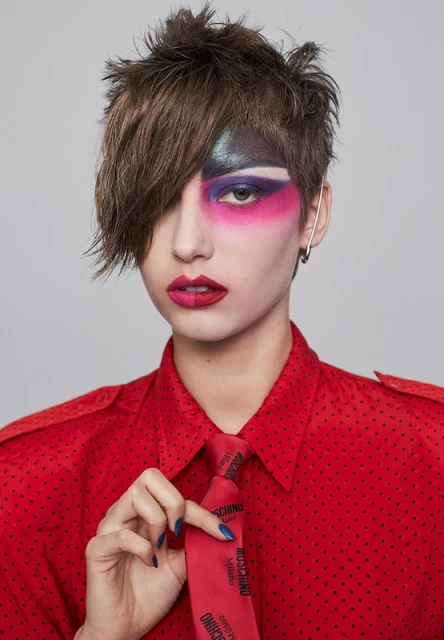 Close-up of a model with half her face in bright makeup, including pink and blue. She is wearing a red shirt and Moschino tie.