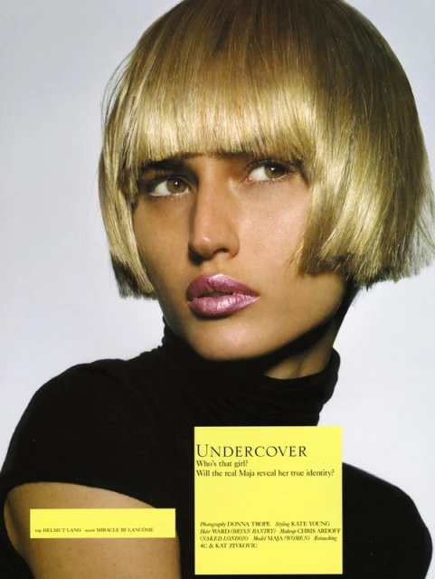 Close-up of a woman with a blunt blonde bob haircut, wearing a black turtleneck, and pink lipstick, against a neutral background in a studio.