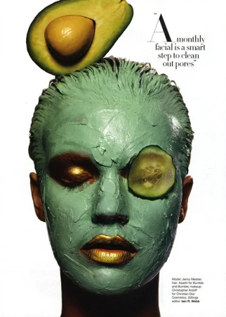 Close-up beauty shot of a woman with a green face mask, avocado on her head, and cucumber on her eye for a skincare theme.