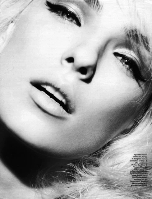 Black and white portrait of a blonde adult female model with heavy eye makeup and slightly open mouth, lying down in photo studio.