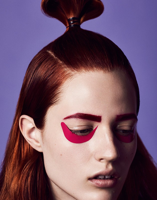 Close-up portrait of a person with creative red hair, graphic makeup, and striking facial features against a violet backdrop.