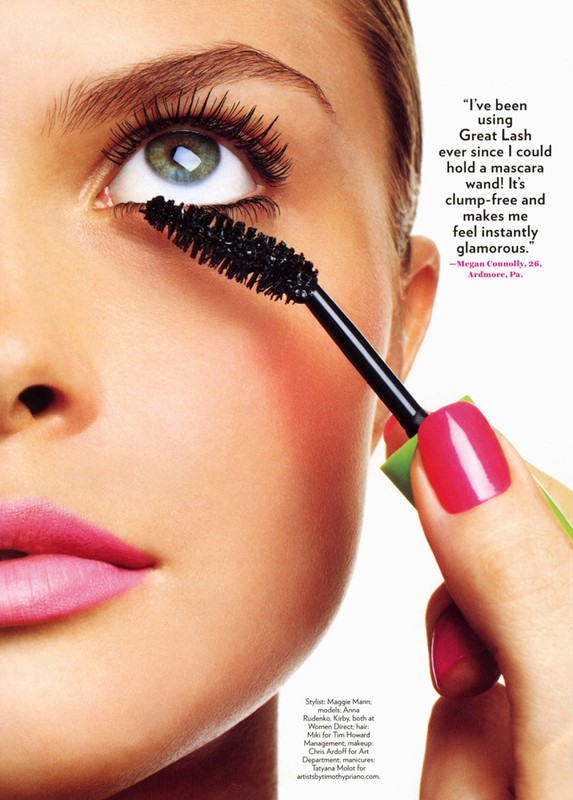Close-up of a woman applying mascara to her lower eyelashes with a black wand, featuring bright pink lipstick and pink fingernail polish.