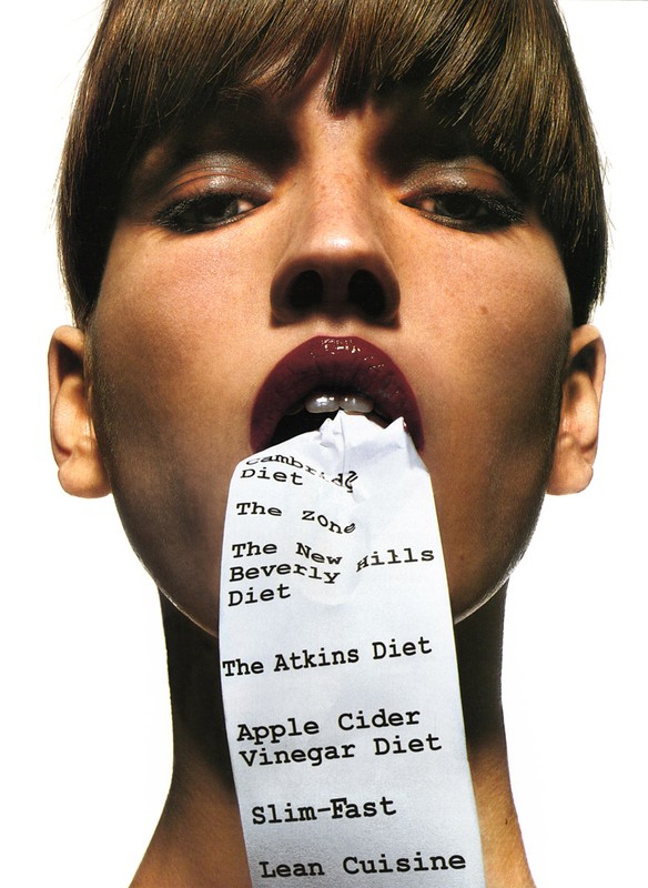 Adult female poses with a crumpled receipt listing various diets in her mouth in a studio shot with bright, even lighting on white.