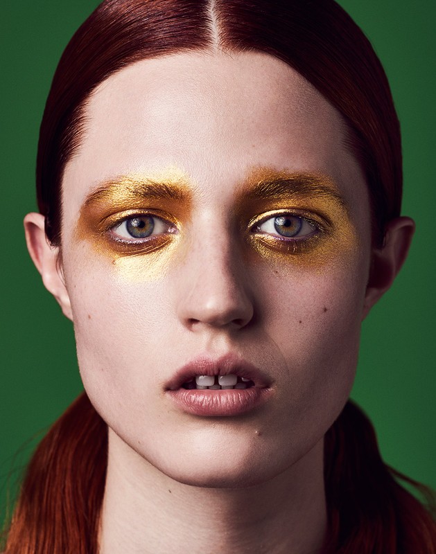 Headshot of a young red-haired woman with golden eye makeup and a green background, emphasizing beauty and high-fashion makeup artistry.