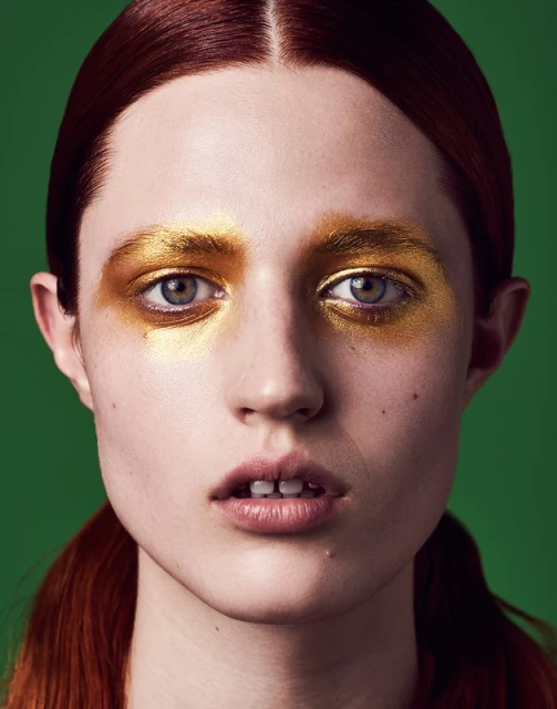 Headshot of a young red-haired woman with golden eye makeup and a green background, emphasizing beauty and high-fashion makeup artistry.