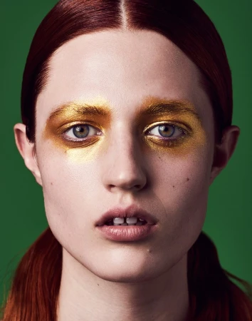 Headshot of a young red-haired woman with golden eye makeup and a green background, emphasizing beauty and high-fashion makeup artistry.