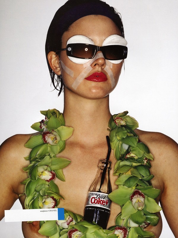 A portrait of a woman with eyewear and bandages, posing with a garland of flowers and a Diet Coke bottle against a white backdrop.