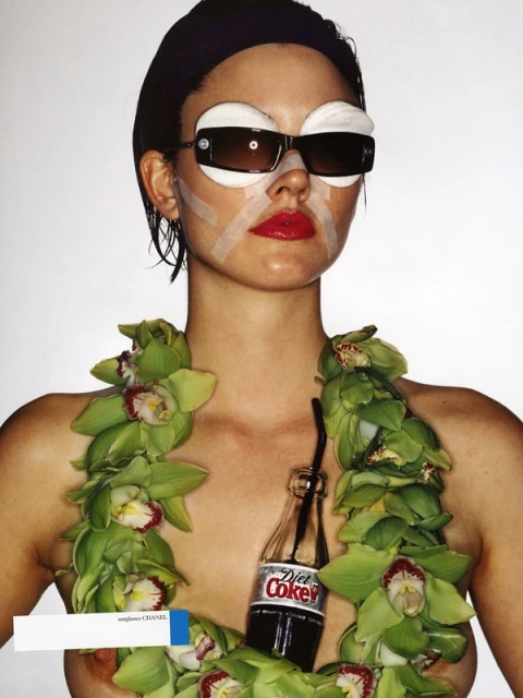 A portrait of a woman with eyewear and bandages, posing with a garland of flowers and a Diet Coke bottle against a white backdrop.