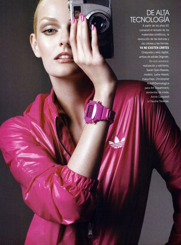 Woman in shiny pink jacket holds camera to eye. Vintage camera and digital watch give modern yet retro tech style. Editorial fashion shot.