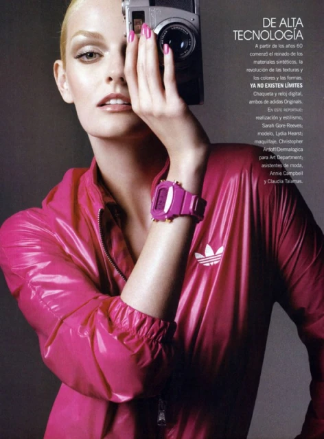 Woman in shiny pink jacket holds camera to eye. Vintage camera and digital watch give modern yet retro tech style. Editorial fashion shot.
