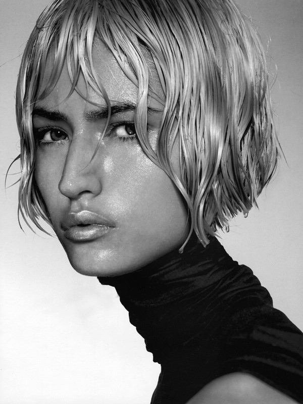 Close-up portrait of a woman with wet styled hair and glossy makeup wearing a dark high-neck top against a grey seamless background