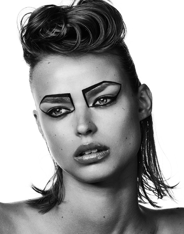 Monochrome portrait of a young adult woman with stylized eye makeup and a wet-look hairstyle against a bright white background.