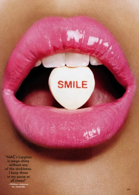Macro shot featuring a woman's pink glossy lips open, revealing a heart-shaped candy with the word 'Smile' written on it.