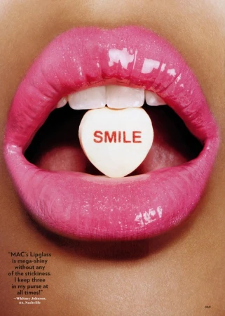 Macro shot featuring a woman's pink glossy lips open, revealing a heart-shaped candy with the word 'Smile' written on it.