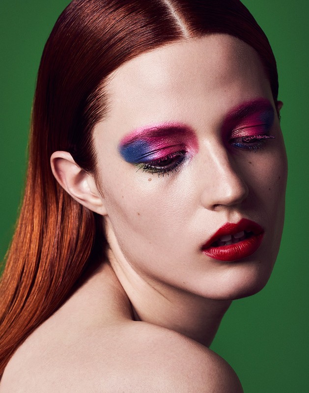 Close-up portrait of an adult female with red hair, bold makeup, including blue and pink eyeshadow, and red lipstick, against a green backdrop.