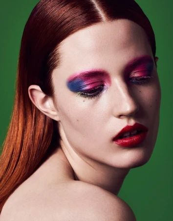Close-up portrait of a woman with red hair and vibrant pink, blue, and green eye makeup against a green background.