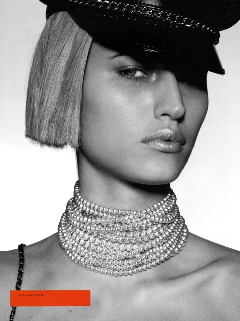 Close-up shot of a woman with blond hair wearing a black hat and large pearl necklace against a white background, fashion editorial.
