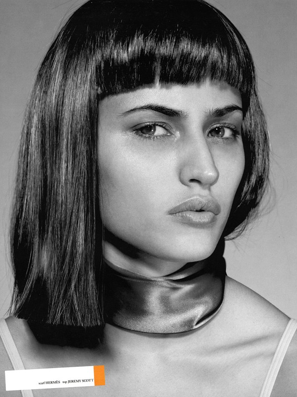 Close-up monochrome portrait of a woman with dark hair, blunt bangs, and a silk scarf around her neck against a grey background.