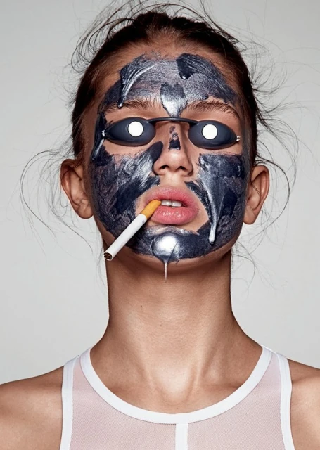 Young woman with a black facial mask, sunglasses, and cigarette in her mouth poses in a studio. Beauty and fashion concept.