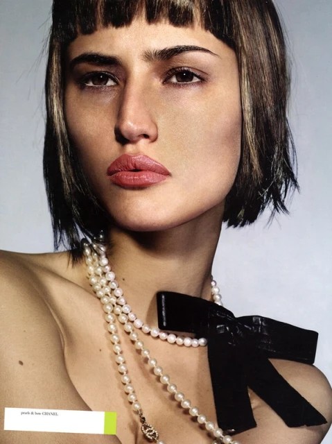 Close-up portrait of a fair-skinned woman with short brown hair and bangs wearing a pearl necklace with a black bow, fashion shot.