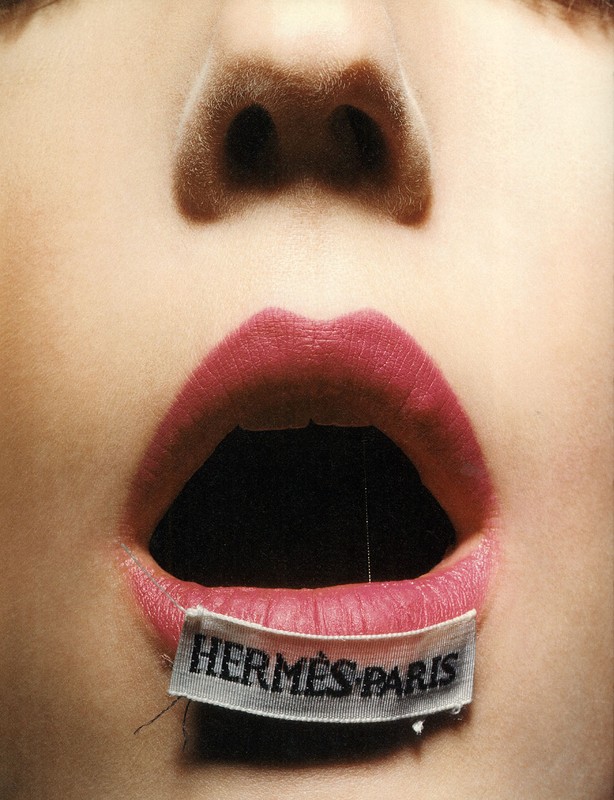 Macro close-up of an adult female's face, showing a woman's open mouth with red lipstick and a Hermes Paris tag on the lower lip.
