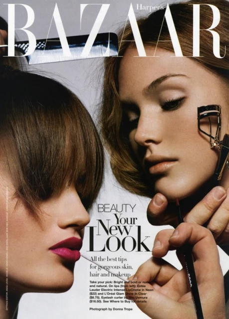 Editorial beauty shot: two fair-skinned adult females having makeup applied, with bright pink lipstick and lash curler, in studio lighting.