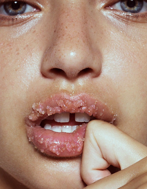 Close-up of a woman's face with sugar scrub on her lips, featuring fair skin, blue eyes, and visible freckles; beauty product shot.