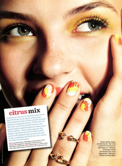 Close-up of a woman's face with colorful flame-designed nail art. She is wearing yellow eyeshadow and gold rings on her fingers.