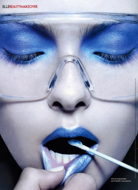 Close-up of a woman's face with blue eyeshadow and lips, holding a cotton swab. She is wearing safety glasses, in a beauty shot.