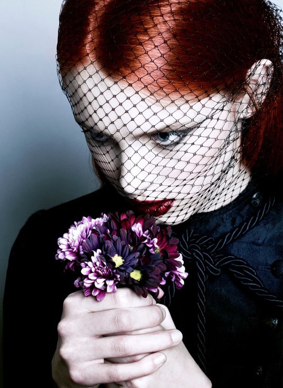 Woman with red hair and dark makeup poses with bouquet, wearing a black veil. Dark and moody fashion beauty shot.