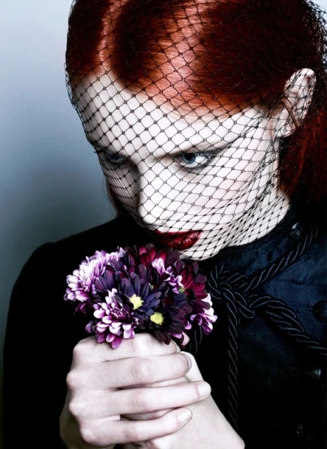 Woman with red hair and dark makeup poses with bouquet, wearing a black veil. Dark and moody fashion beauty shot.