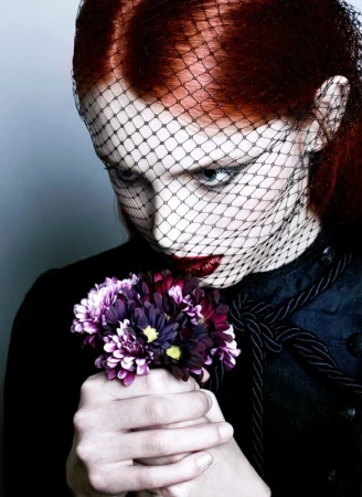 Woman with red hair and dark makeup poses with bouquet, wearing a black veil. Dark and moody fashion beauty shot.