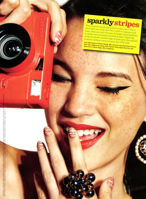 Close-up of a smiling woman holding a red camera near her face, featuring unique nail art, a black ring, and statement earrings.