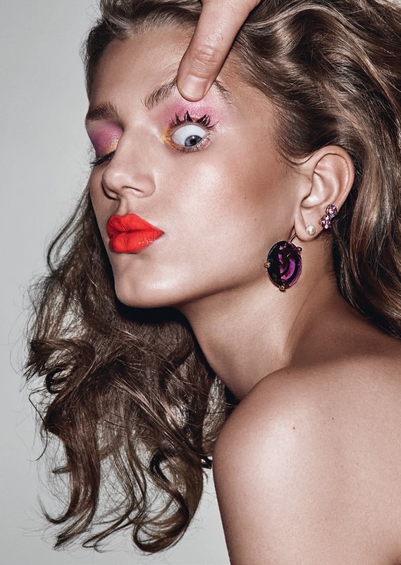 Close-up of a fair-skinned adult female model with red lipstick, pink eye shadow, and wavy brunette hair with a hand pulling her eye open.