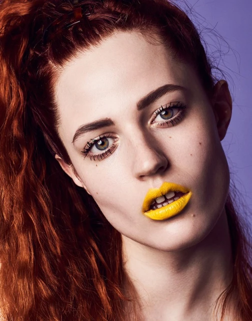 Close-up portrait of a young woman with auburn hair and bright yellow lipstick against a solid light purple background.