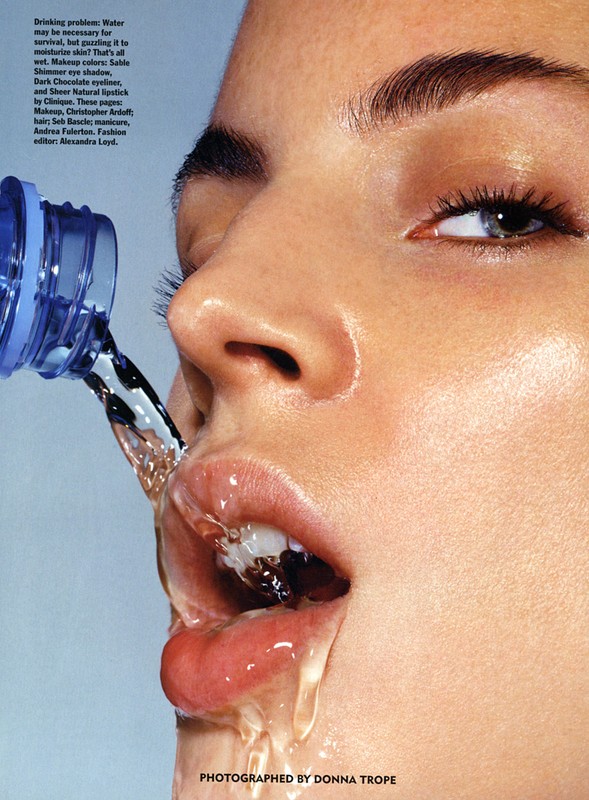 Close-up of an adult female face being splashed with water from a blue plastic bottle, highlighting skincare and hydration.
