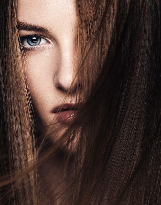 Close-up portrait of a woman with striking blue eyes, her face partially covered by flowing brunette hair in a beauty shot.