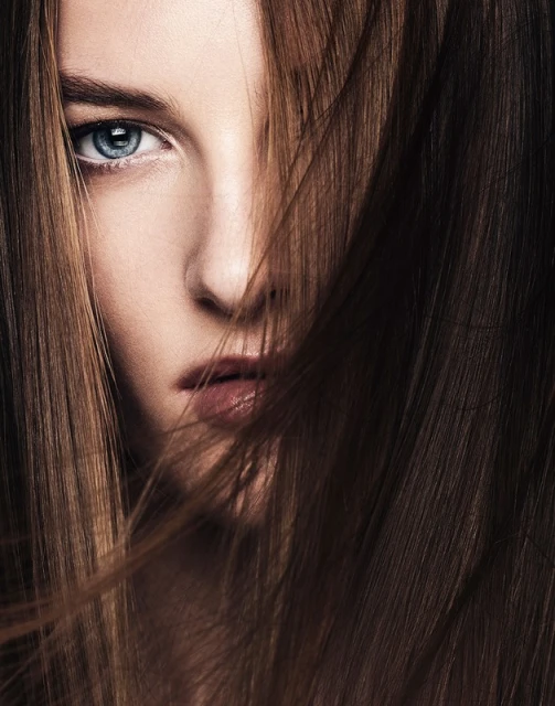 Close-up portrait of a woman with striking blue eyes, her face partially covered by flowing brunette hair in a beauty shot.