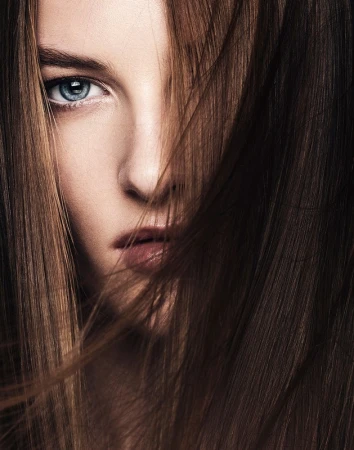 Close-up portrait of a woman with striking blue eyes, her face partially covered by flowing brunette hair in a beauty shot.