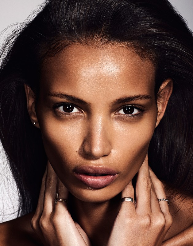 Close-up portrait of a Black woman with dark hair and fair skin wearing rings, gazing directly at the camera against a light background.
