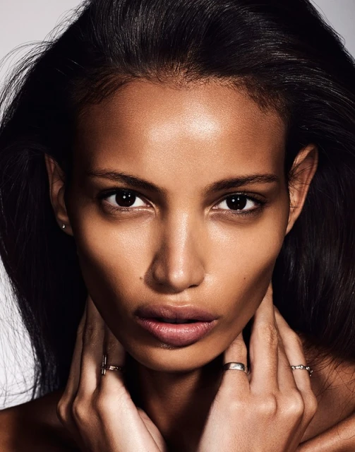 Close-up portrait of a Black woman with dark hair and fair skin wearing rings, gazing directly at the camera against a light background.