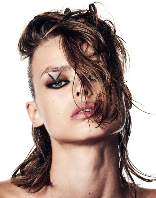 Close-up portrait of a woman with slicked-back hair, dark eye makeup, and a cross shape near her eye against a bright white background.