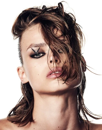 Close-up portrait of a woman with slicked-back hair, dark eye makeup, and a cross shape near her eye against a bright white background.